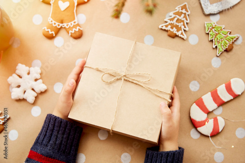 Child holds gift box. Crafting decoration, gingerbread cookies, branch of fir tree, hot beverage with marshmallows and Christmas ornaments. Trendy DIY style. Merry Christmas concept photo