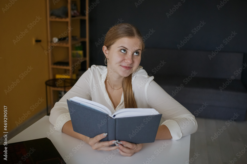 A young successful employee of a business woman in a white shirt holds a blue notepad in her hands and looks to the side, sits at a white desk workplace A young successful employee of a business woman in a white shirt holds a blue notepad in her hands and looks to the side, sits at a white desk workplace