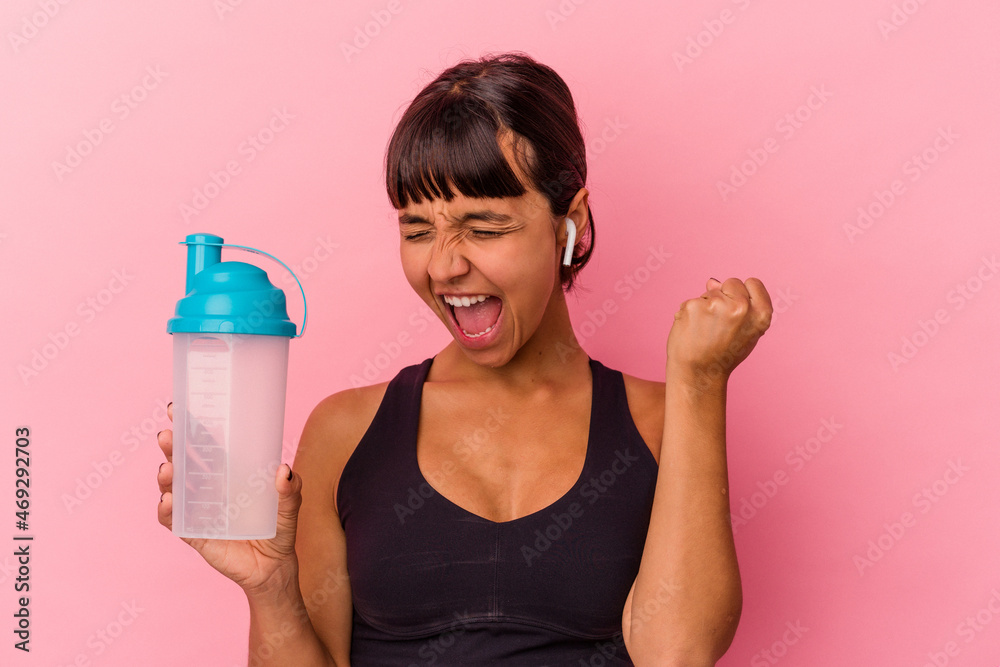 Young mixed race woman drinking protein shake isolated on pink background raising fist after a victory, winner concept. Young mixed race woman drinking protein shake isolated on pink background raising fist after a victory, winner concept.
