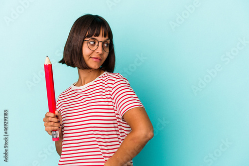 Young mixed race woman holding a big pencil isolated on blue background looks aside smiling  cheerful and pleasant.