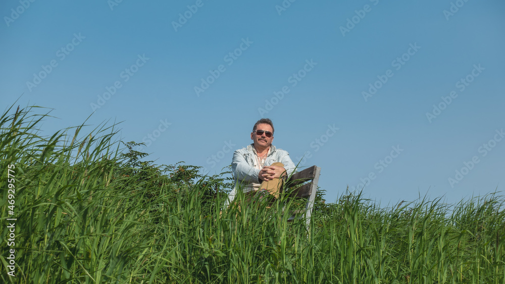 A man in dark glasses is sitting resting on a bench among the lush tall green grass. Legs crossed. There's a smile on his face. In the background is a clear blue sky. A sunny summer day. Kamchatka A man in dark glasses is sitting resting on a bench among the lush tall green grass. Legs crossed. There's a smile on his face. In the background is a clear blue sky. A sunny summer day. Kamchatka
