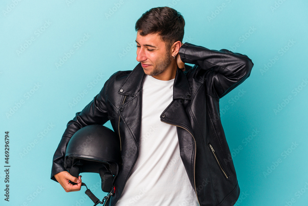 Young biker man holding helmet isolated on blue background touching back of head, thinking and making a choice. Young biker man holding helmet isolated on blue background touching back of head, thinking and making a choice.