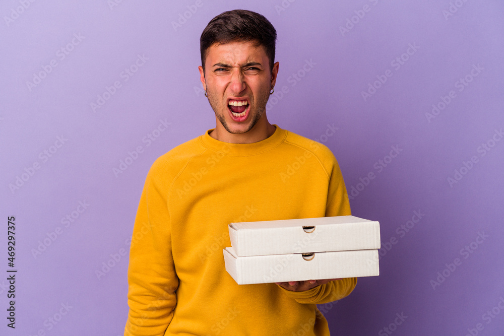 Young caucasian man holding pizzas isolated on purple background screaming very angry and aggressive. Young caucasian man holding pizzas isolated on purple background screaming very angry and aggressive.