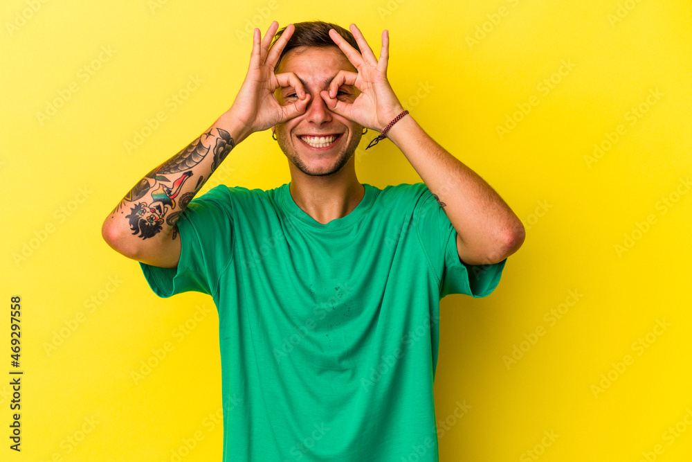 Young caucasian man with tattoos isolated on yellow background showing okay sign over eyes Young caucasian man with tattoos isolated on yellow background showing okay sign over eyes