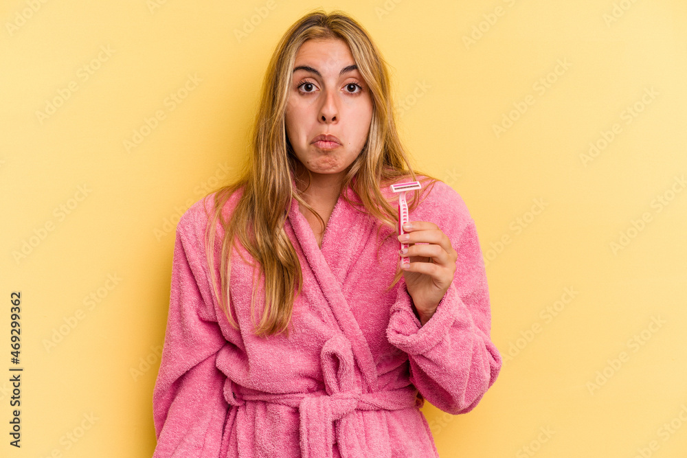 Young caucasian blonde woman wearing bathrobe holding blades isolated on yellow background shrugs shoulders and open eyes confused. Young caucasian blonde woman wearing bathrobe holding blades isolated on yellow background shrugs shoulders and open eyes confused.