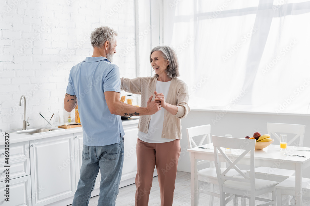 Positive woman holding hand of tattooed husband near fruits on table in kitchen. Positive woman holding hand of tattooed husband near fruits on table in kitchen.