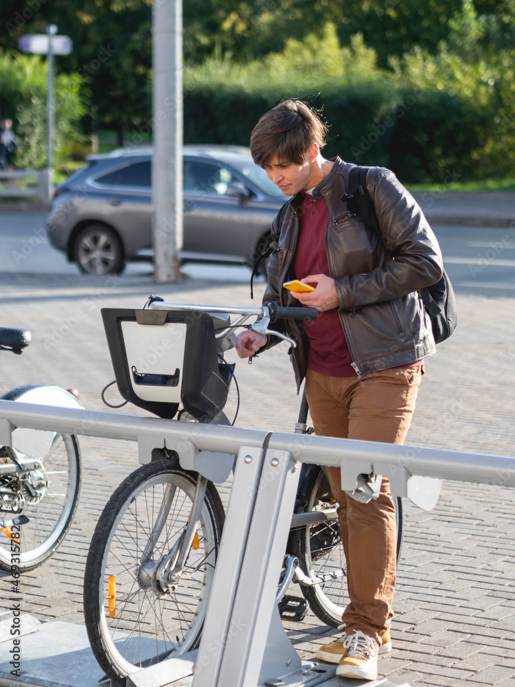 Obraz premium Young man is renting bicycle by smartphone application. Eco-friendly urban transport for rent. Healthy lifestyle in modern town. Autumn sunset.