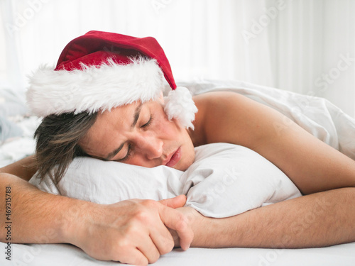 Man in red Santa hat is sleeping after Christmas or New Year celebration. Early morning in bedroom lit with light. © Konstantin Aksenov