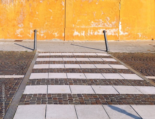 Crosswalk made of white stone on porphyry street with metal poles at its end. Peeled and weathered yellow wall on behind. Background for copy space.