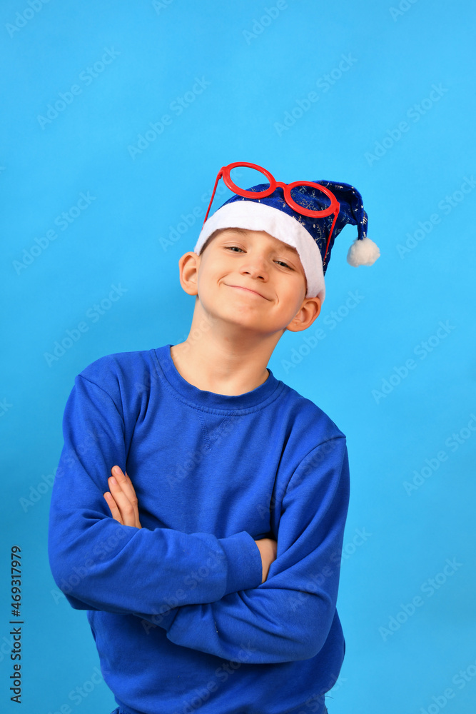 A joyful and smiling boy in a blue Santa Claus hat and red glasses on his head crossed his arms and poses for New Year and Christmas. A joyful and smiling boy in a blue Santa Claus hat and red glasses on his head crossed his arms and poses for New Year and Christmas.