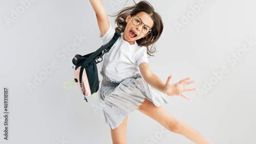 Horizontal studio image of a positive schoolgirl in eyeglasses with backpack smiling and jumping feels happy. Schoolkid has a joyful expression posing over a studio grey background.