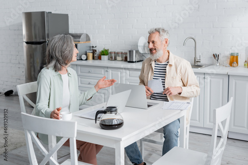 Smiling man holding bills near wife and laptops in kitchen.