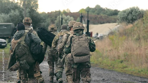 A group of military men returning after completing a task. Soldiers in military uniform with machine guns walk on a stony road with their backs to the cell. photo