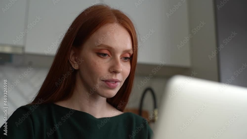 Close-up low-angle view of attractive smiling young woman using typing on laptop computer sitting at desk in light room. Tracking shot of pretty redhead female freelancer working at home office. Close-up low-angle view of attractive smiling young woman using typing on laptop computer sitting at desk in light room. Tracking shot of pretty redhead female freelancer working at home office.