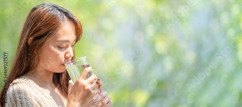 Asian Woman drinks water from tall glass of water at outdoor tree bokeh green for banner background.