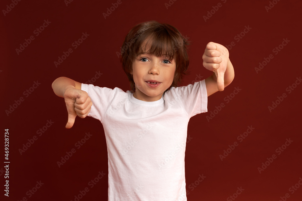 Indoor image of small Caucasian brunette boy showing his attitude putting thumbs down, smiling, dressed in white, having cute stylish haircut. Human emotions, feelings, body language, signs Indoor image of small Caucasian brunette boy showing his attitude putting thumbs down, smiling, dressed in white, having cute stylish haircut. Human emotions, feelings, body language, signs