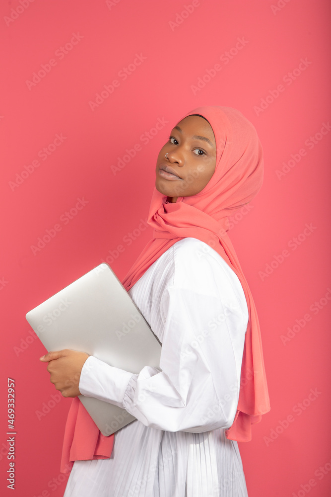A young African American woman in profile wearing pink scarf and holding a laptop isolated on pink background