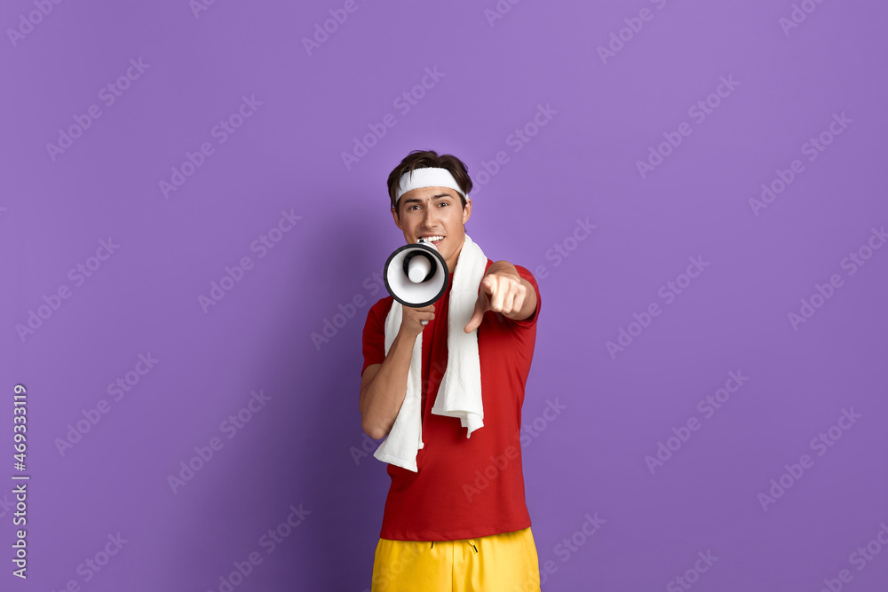 Obraz premium Hey you, start today. Young sportsman is holding loudspeaker and points to camera with finger, wears red t-shirt, yellow shorts, white headband and towel over neck, isolated on violet background.