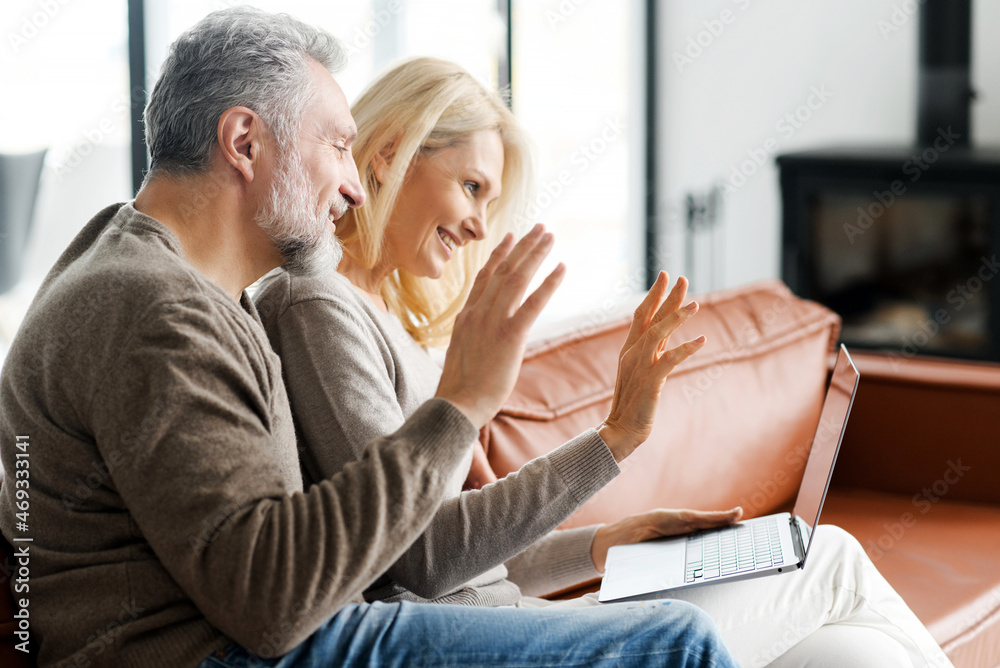 Positive middle-aged couple is using laptop for video call sitting on comfortable sofa in cozy living room. Cheerful spouses waving into webcam. Online communication concept
