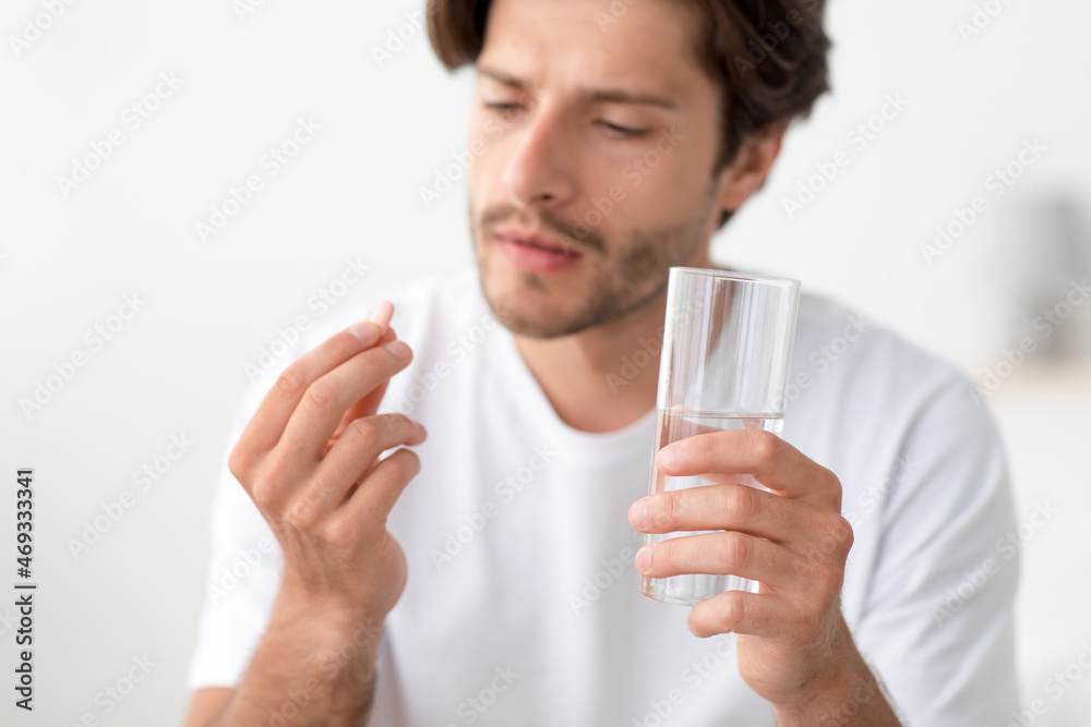 Pensive millennial european man hold glass of water and look at pill in hand Pensive millennial european man hold glass of water and look at pill in hand