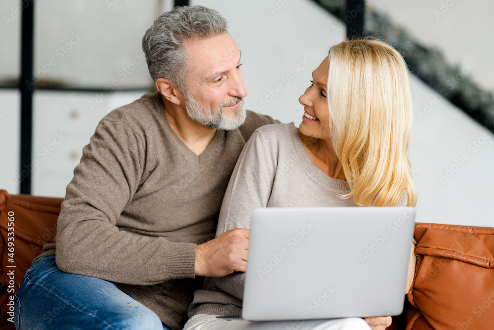 Middle-aged couple sitting on the couch and using laptop for spending leisure together, watching movies, web surfing, shopping online. Cheerful senior spouses with a laptop at home look at each other Middle-aged couple sitting on the couch and using laptop for spending leisure together, watching movies, web surfing, shopping online. Cheerful senior spouses with a laptop at home look at each other
