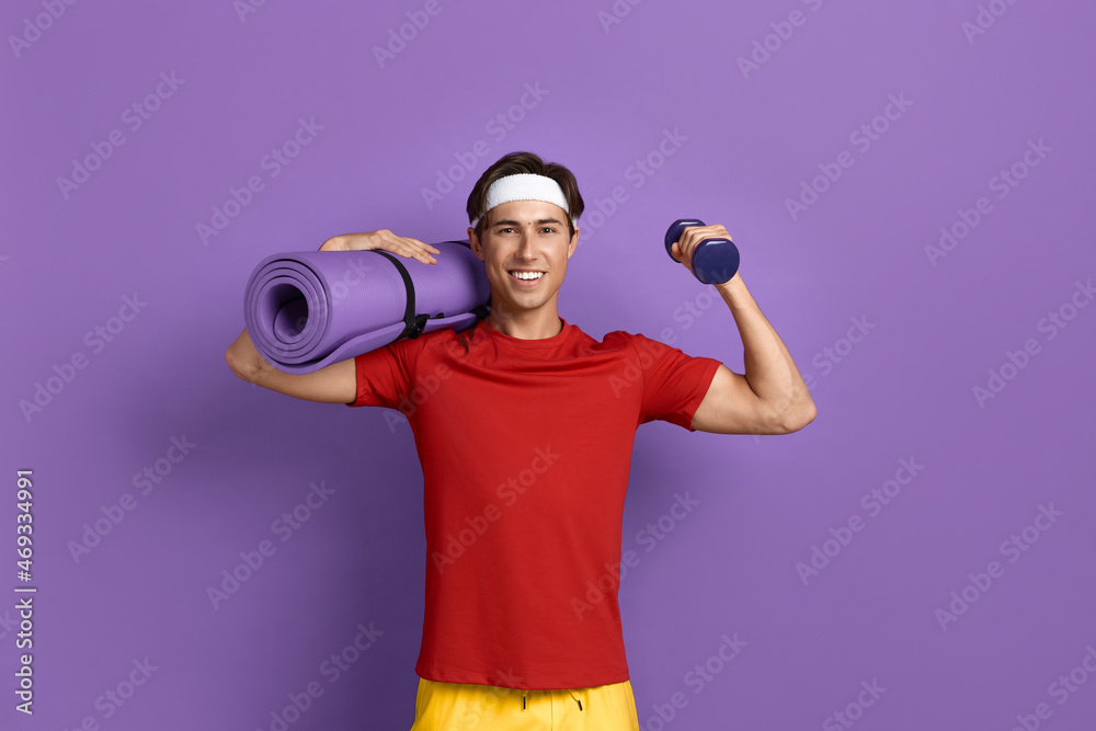 Smiling handsome athlete holds fitness or yoga mat on shoulder and raises arm with dumbbell, shows muscles, biceps, wears red t-shirt, yellow shorts and white headband, isolated on violet background.