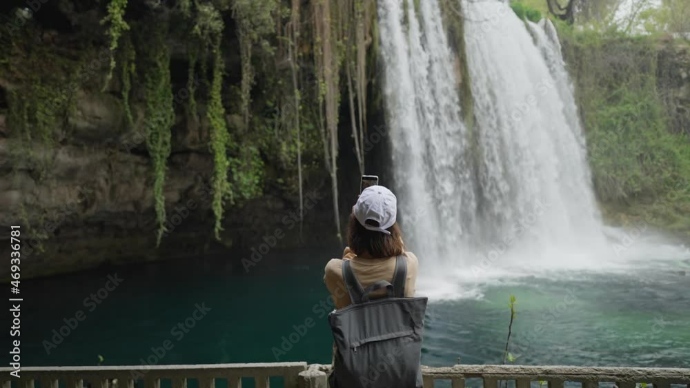 rear view slow motion camera zoming out of woman tourist taking photo or video on mobile phone on huge Waterfall background in Antalya. Famous view point and touristic place in Turkey