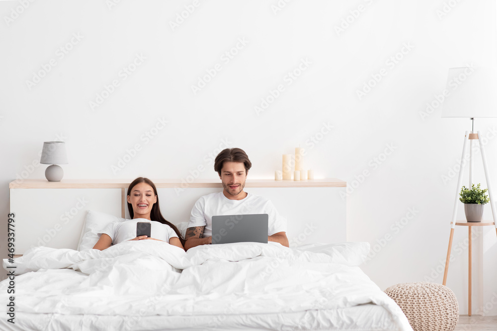 Cheerful european young wife typing on phone, husband surfing in internet on laptop in bed Cheerful european young wife typing on phone, husband surfing in internet on laptop in bed