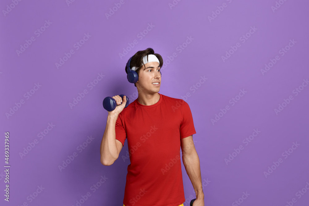 Envious young sportsman is raising arm with dumbbell, looks aside at muscles of gym rat, wants quick result, wears red t-shirt, blue headphones and white headband, isolated on violet background. Envious young sportsman is raising arm with dumbbell, looks aside at muscles of gym rat, wants quick result, wears red t-shirt, blue headphones and white headband, isolated on violet background.