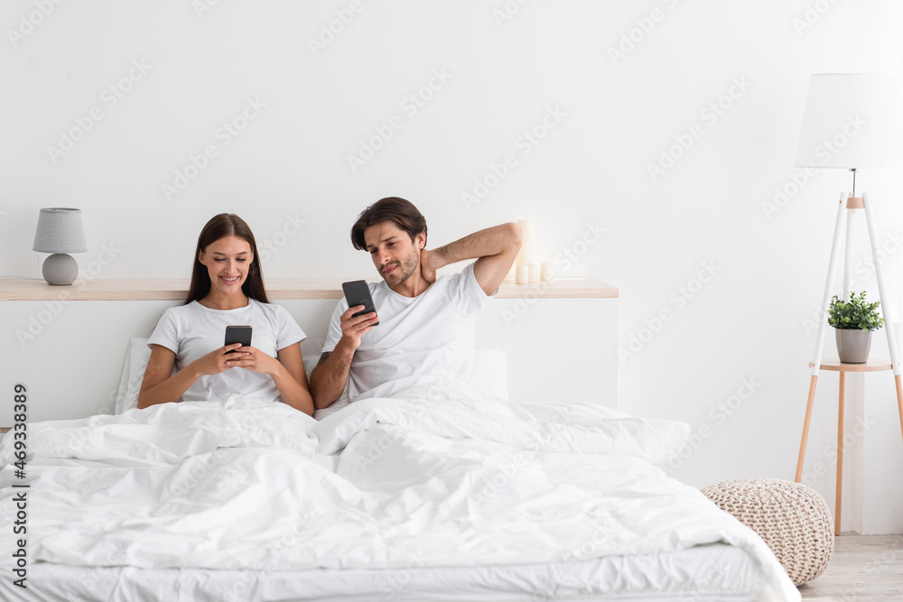 Attractive millennial european husband and wife read news and chatting on phones in morning in bed Attractive millennial european husband and wife read news and chatting on phones in morning in bed