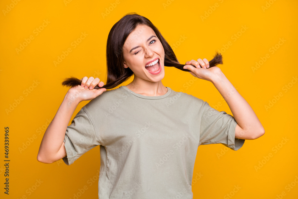 Photo of funky young pretty lady wink eye hold hands hair wear t-shirt isolated on yellow color background Photo of funky young pretty lady wink eye hold hands hair wear t-shirt isolated on yellow color background