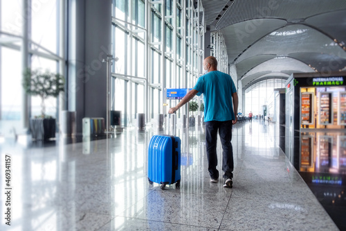 Man pulling suitcase in modern airport terminal. Travelling guy wearing casual clothes and mask walking away with his luggage while waiting for transport. Copy space