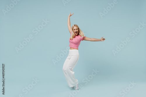 Full body smiling young blonde woman 20s wearing casual pink t-shirt leaning back stand on toes fooling around isolated on plain pastel light blue background studio portrait. People lifestyle concept.