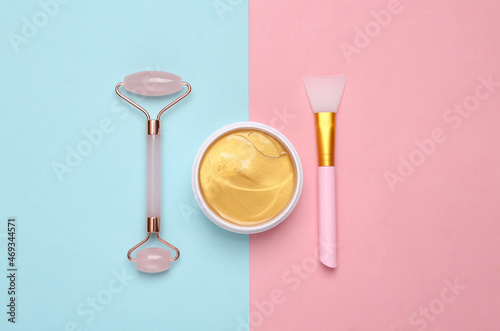 Gold cream jar or patches, facial massage roller on pink blue background. Skin care, lifting face concept