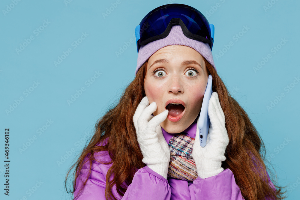 Fototapeta premium Skier scared astonished woman in warm purple padded windbreaker jacket ski goggles mask spend extreme weekend in mountains talk on mobile cell phone hold face isolated on plain blue background studio