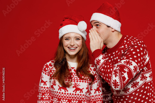 Young happy couple friends two man woman in sweater hat whispering gossip and tell secret behind his hand share news hold face isolated on plain red background Happy New Year 2022 celebration concept.