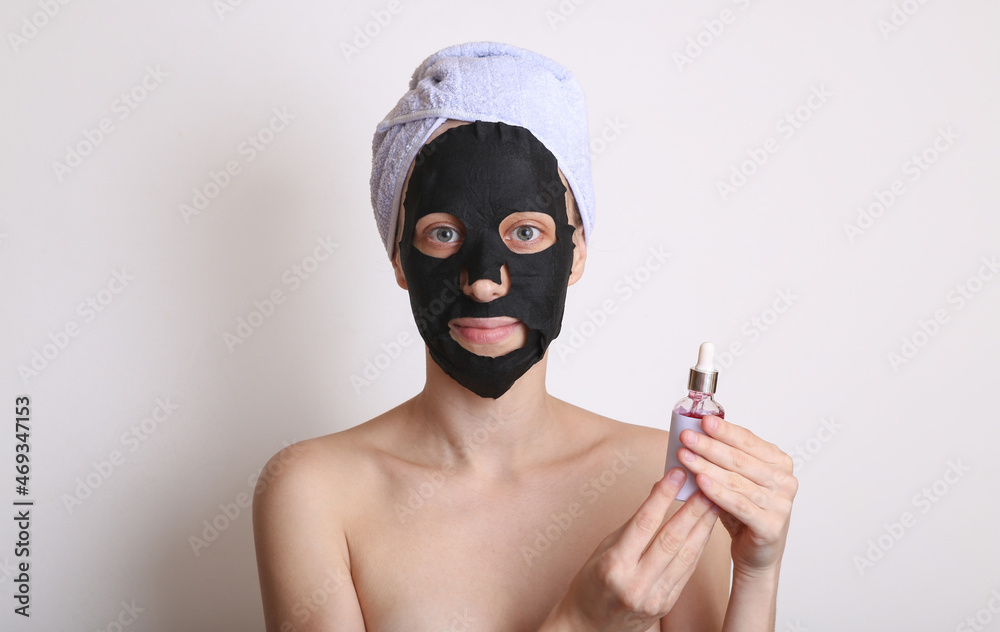 Beautiful woman with purifying black mask on her face and towel on head. Girl holding bottle of serum. White background. Facial skin care Beautiful woman with purifying black mask on her face and towel on head. Girl holding bottle of serum. White background. Facial skin care