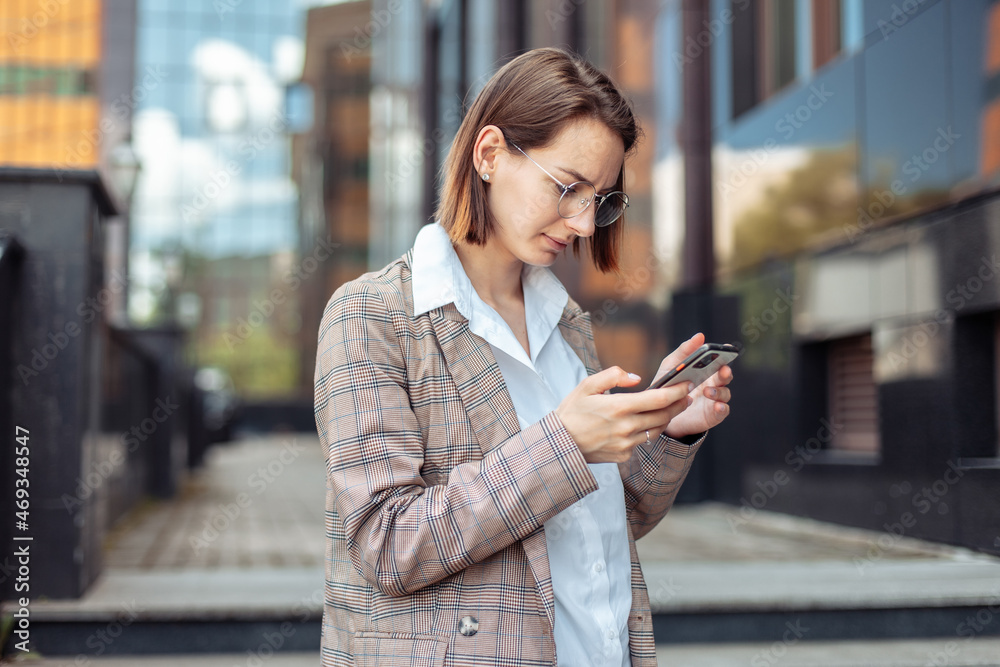 Fototapeta premium Young modern business woman in a jacket and glasses looking at the screen of a smartphone in the city