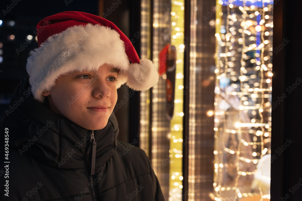 Boy in Santas hat looking and dreaming in illuminated shop window. Xmas presents holidays, or shopping on New Year or Christmas concept
