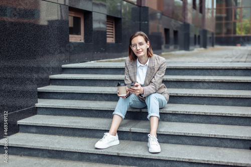 Young stylish businesswoman sitting on the steps and holding a phone and a cup of coffee