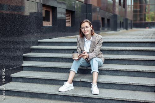 Young stylish businesswoman sitting on the steps and holding a phone and a cup of coffee © splitov27