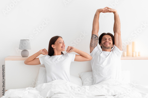 Happy caucasian millennial wife and husband woke up and stretching body sitting on bed in white bedroom #469349371