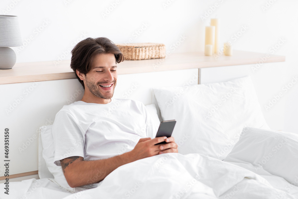 Happy european millennial man relax, typing on phone, sitting in bed in white bedroom Happy european millennial man relax, typing on phone, sitting in bed in white bedroom