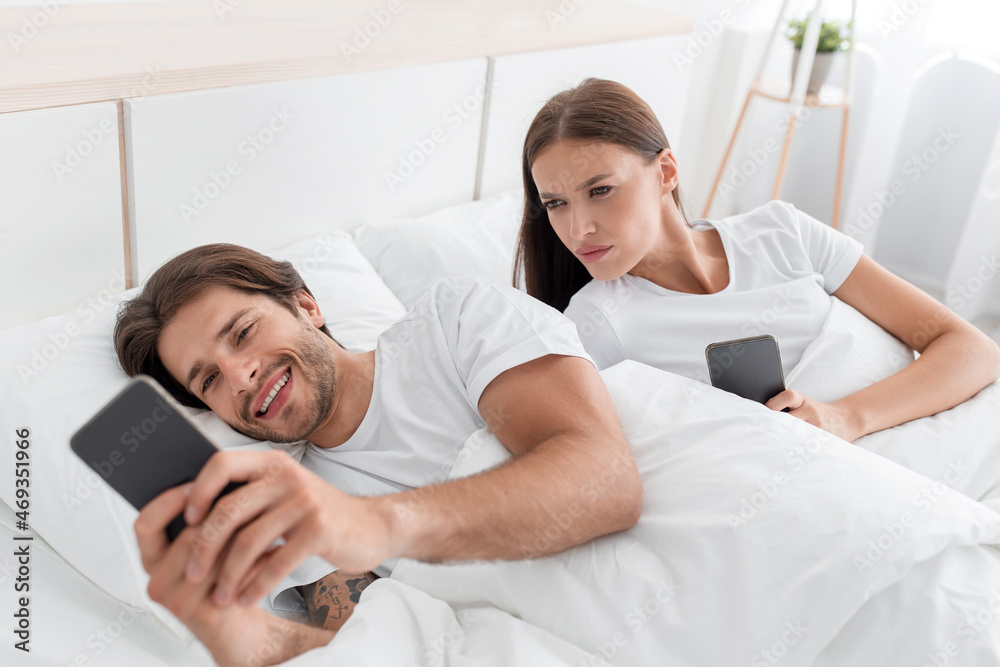 Offended angry young european wife looks at husbands phone and jealous, on bed in bedroom interior