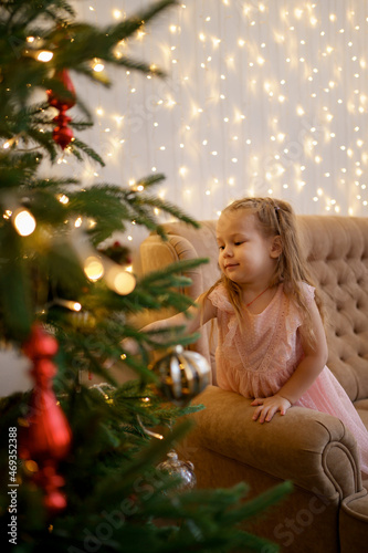 Little girl looking to a fir tree on Christmas tree making a wish while standing on sofa at home with lights in background