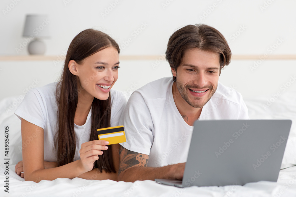 Happy millennial caucasian woman and guy lie on bed paying for purchase, order with laptop and credit card Happy millennial caucasian woman and guy lie on bed paying for purchase, order with laptop and credit card