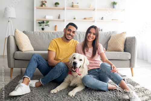 Full length portrait of positive international couple with their pet dog sitting on soft carpet at home photo