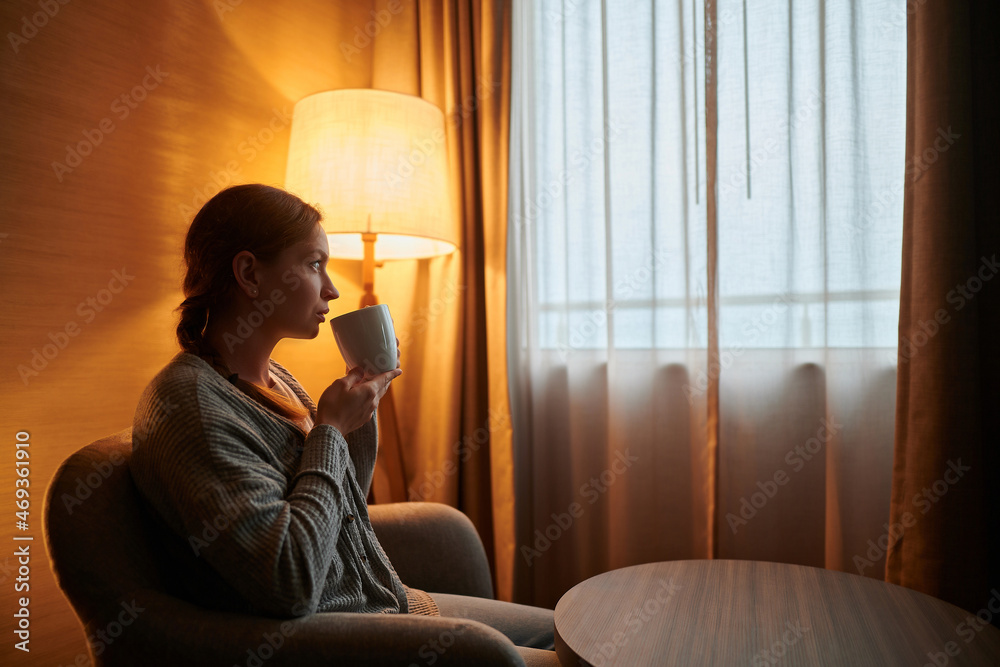 A young blonde woman in home clothes sits near the window with a cup in her hand. Soft light from a floor lamp. Rest, relaxation, hotel, home comfort, quarantine, self-isolation. A young blonde woman in home clothes sits near the window with a cup in her hand. Soft light from a floor lamp. Rest, relaxation, hotel, home comfort, quarantine, self-isolation.