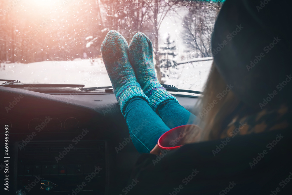 Young slender woman has thrown her feet in warm socks on the dashboard of the car and is holding a cup of hot tea in her hands. Concept of travel, trips in winter. - obrazy, fototapety, plakaty 