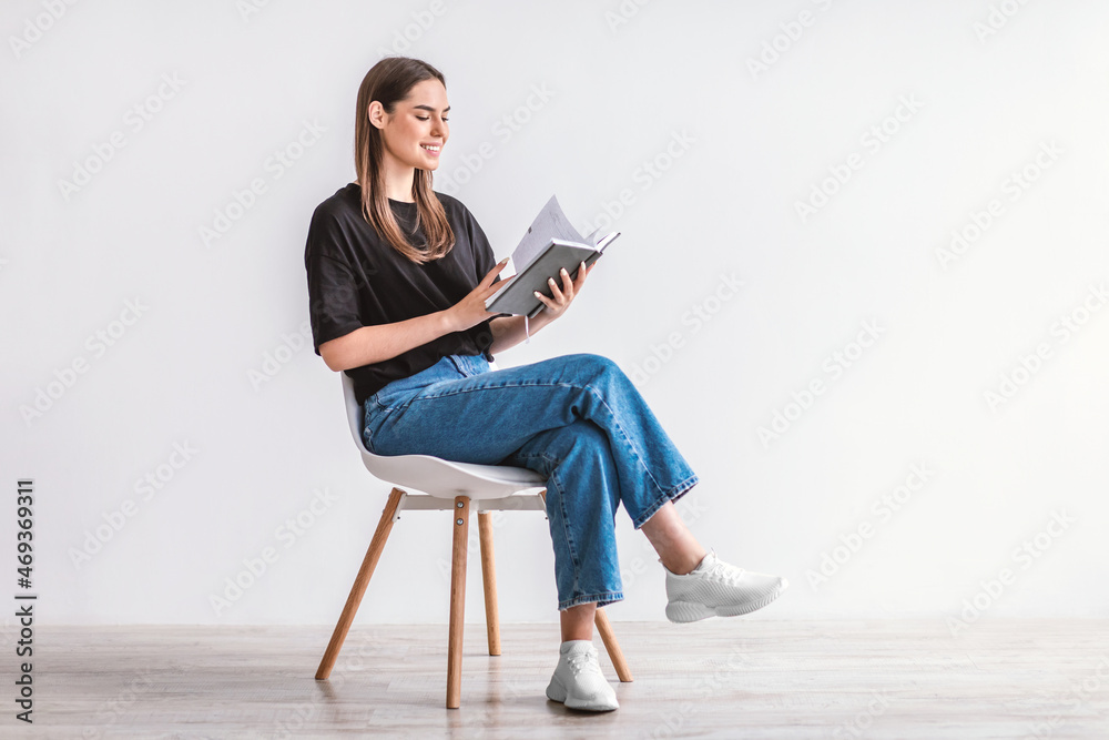 Happy young Caucasian woman reading book, sitting on chair against white studio wall, copy space